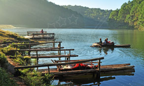 Jangadas de bambu atracadas ao longo da margem de um lago enevoado, com pessoas a remar uma jangada junto a colinas arborizadas ao amanhecer