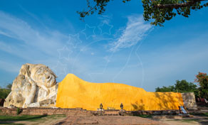 Grande estátua de Buda reclinado coberta com tecido amarelo vivo sob céu azul limpo, enquadrada por ramos de árvore