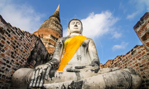 Estátua de pedra de Buda sentado com faixa laranja entre ruínas de templo em tijolo com um chedi dourado sob céu azul