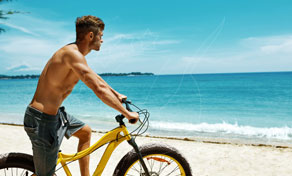 Homem sem camisa de bermuda jeans empurra bicicleta beach cruiser amarela pela areia branca junto ao mar turquesa