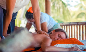 Mulher deitada de bruços sobre toalha laranja recebe massagem nas costas em pavilhão de spa de bambu ao ar livre