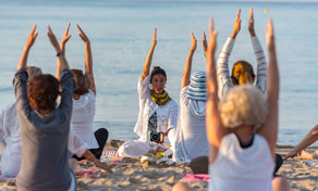 Grupo sentado na areia com os braços erguidos numa aula de yoga guiada por instrutor de frente para o mar