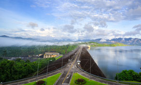 Estrada no coroamento de uma grande barragem junto a uma albufeira calma, com montanhas calcárias enevoadas ao amanhecer