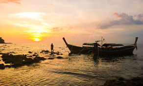 Barcos longtail ancorados em água rasa ao pôr do sol com silhueta de pessoa caminhando perto da costa rochosa