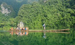 Jangada de bambu com turistas a atravessar rio verde calmo sob colinas de karst calcário arborizadas