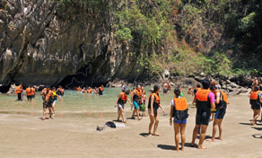 Turistas de coletes salva-vidas laranja numa praia arenosa dentro de lagoa calcária rodeada de falésias com selva