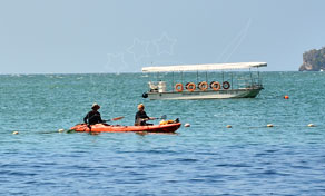 Dois remadores num caiaque laranja em água turquesa calma a passar por barco turístico com toldo perto de um cabo