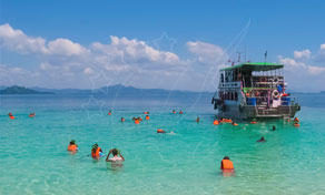 Mergulhadores de snorkel com coletes salva-vidas laranja nadam em águas turquesa rasas junto a um barco de excursão de madeira perto de uma ilha tropical