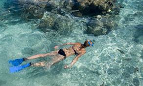 Dois mergulhadores de snorkel com nadadeiras azuis flutuam de bruços em água turquesa rasa sobre corais