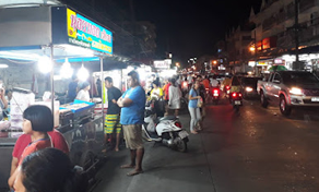 Movimentado mercado noturno de rua com bancas de comida iluminadas, motos estacionadas e uma pick-up branca a passar