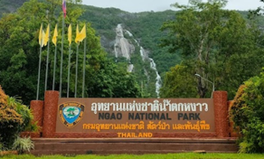 Placa de entrada do Parque Nacional de Ngao, Tailândia, com bandeiras amarelas e uma cascata alta na falésia florestada ao fundo