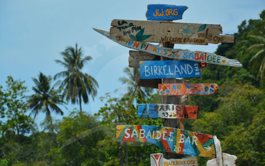 Placa de madeira pintada à mão com setas de destinos junto a palmeiras e selva sob céu azul