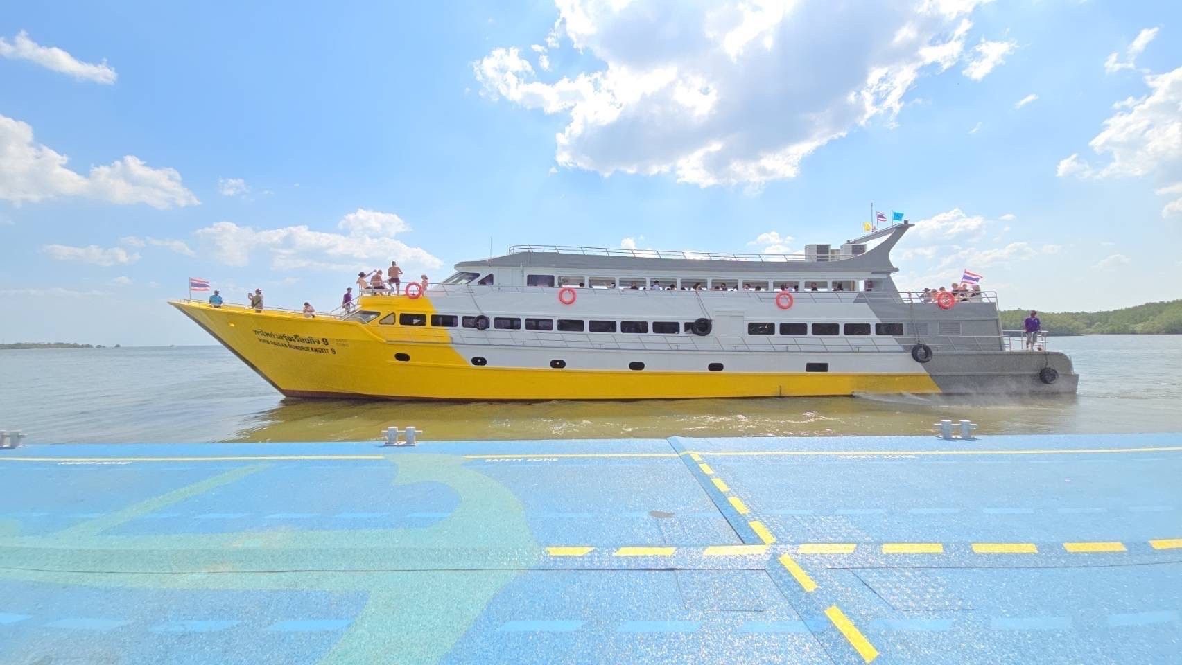 Ferry de passageiros amarelo e branco com turistas no deck a afastar-se de um cais de betão azul sob nuvens luminosas