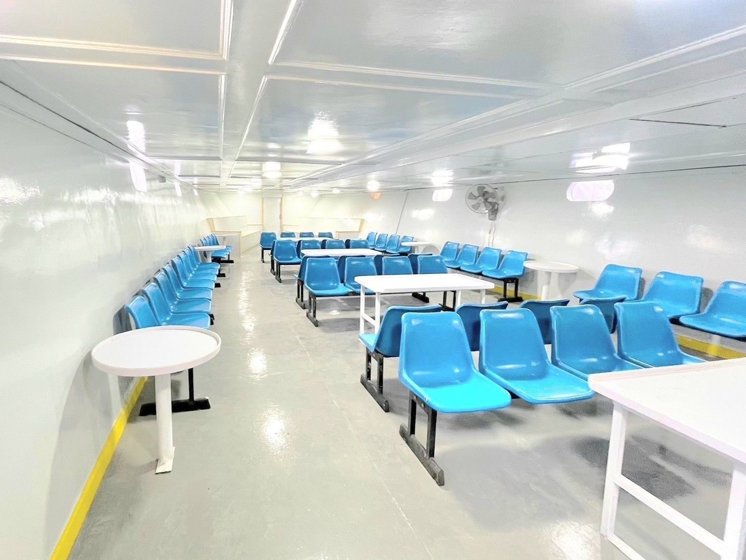 Cabina de passageiros vazia de um ferry com filas de assentos azuis de plástico, mesas brancas e teto branco iluminado