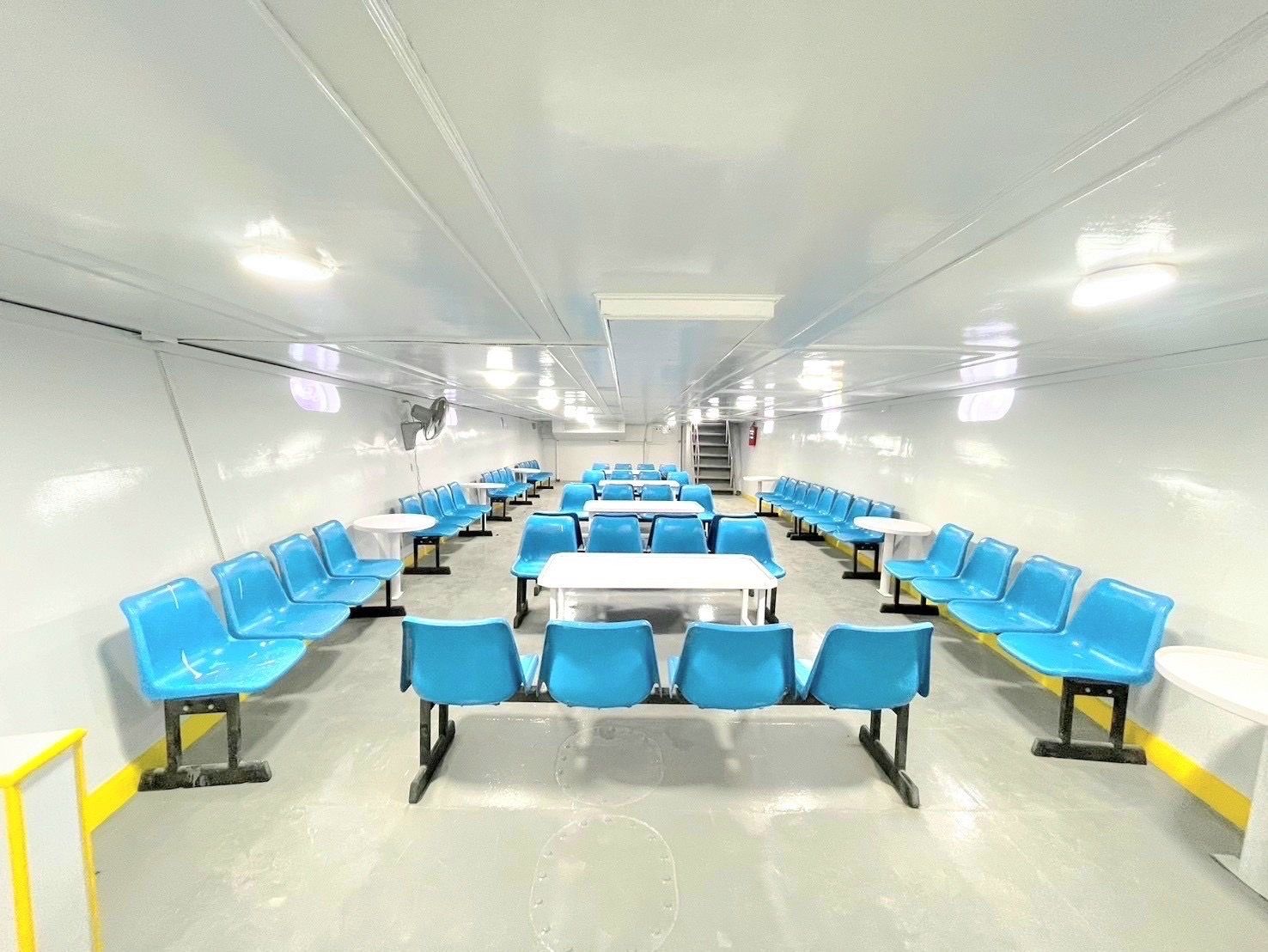 Cabina de passageiros vazia de um ferry com filas de bancos azuis de plástico, mesas brancas e interior branco luminoso