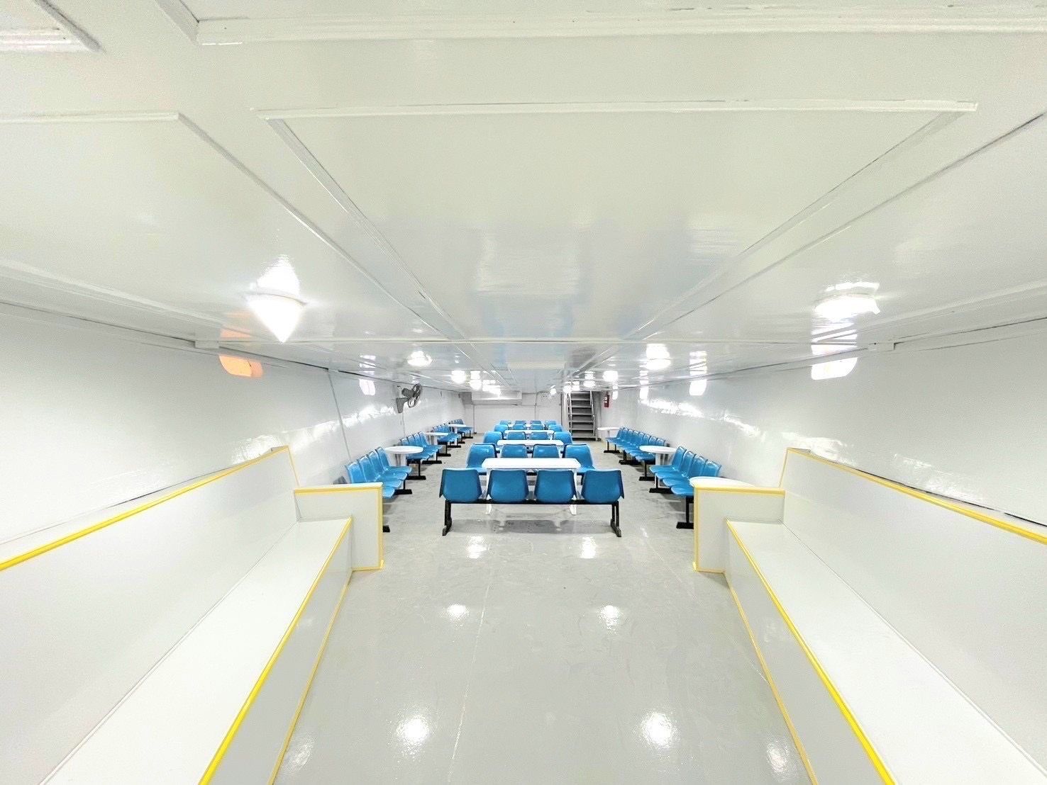 Cabina de passageiros vazia de um ferry com filas de bancos azuis, mesas brancas e paredes e chão brancos brilhantes