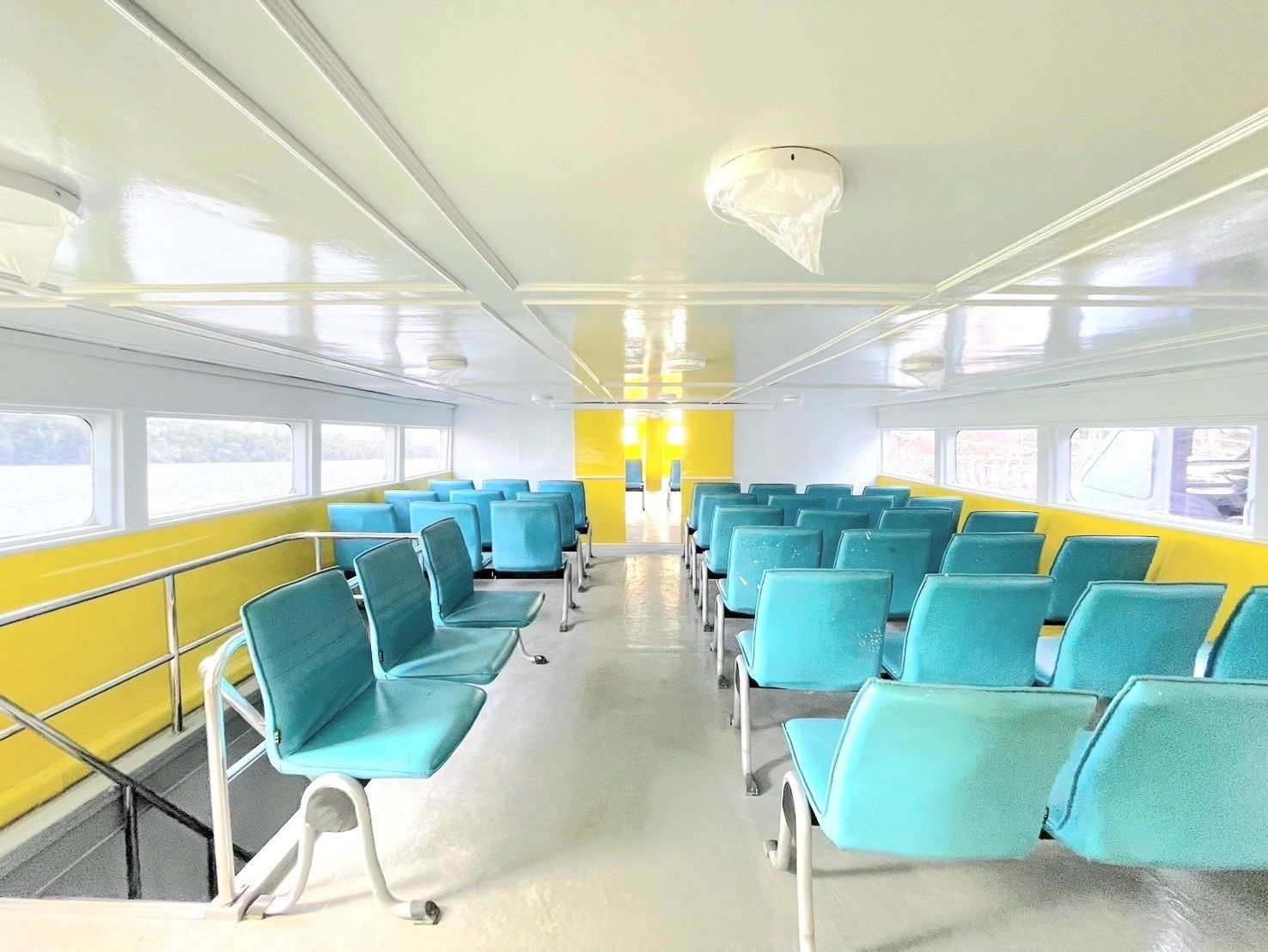 Cabina de passageiros vazia de um ferry com filas de assentos turquesa, paredes amarelas e janelas com vista para o mar e a costa