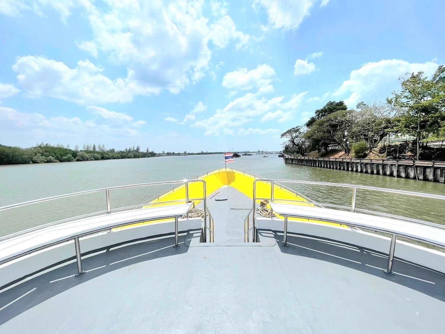Deck de proa de um ferry de passageiros amarelo e cinzento a navegar num rio largo junto a uma margem de mangal e um cais