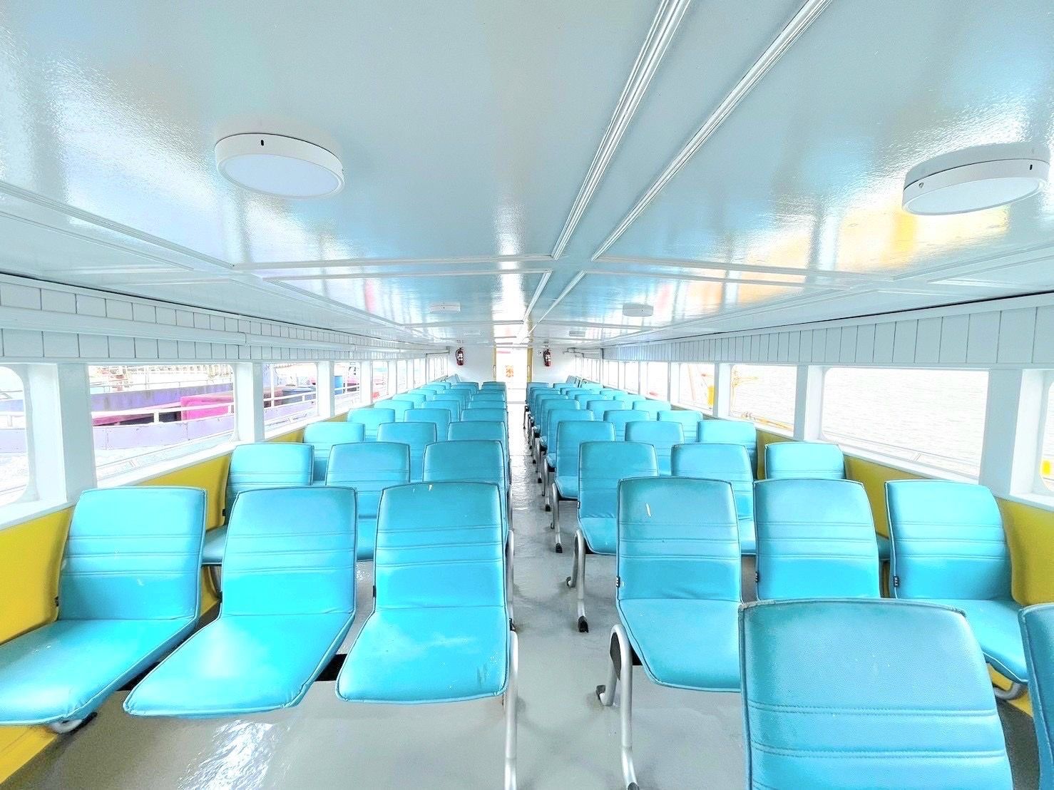 Cabina de passageiros vazia de um ferry com filas de assentos reclináveis de vinil turquesa, teto branco e janelas com vista para o cais