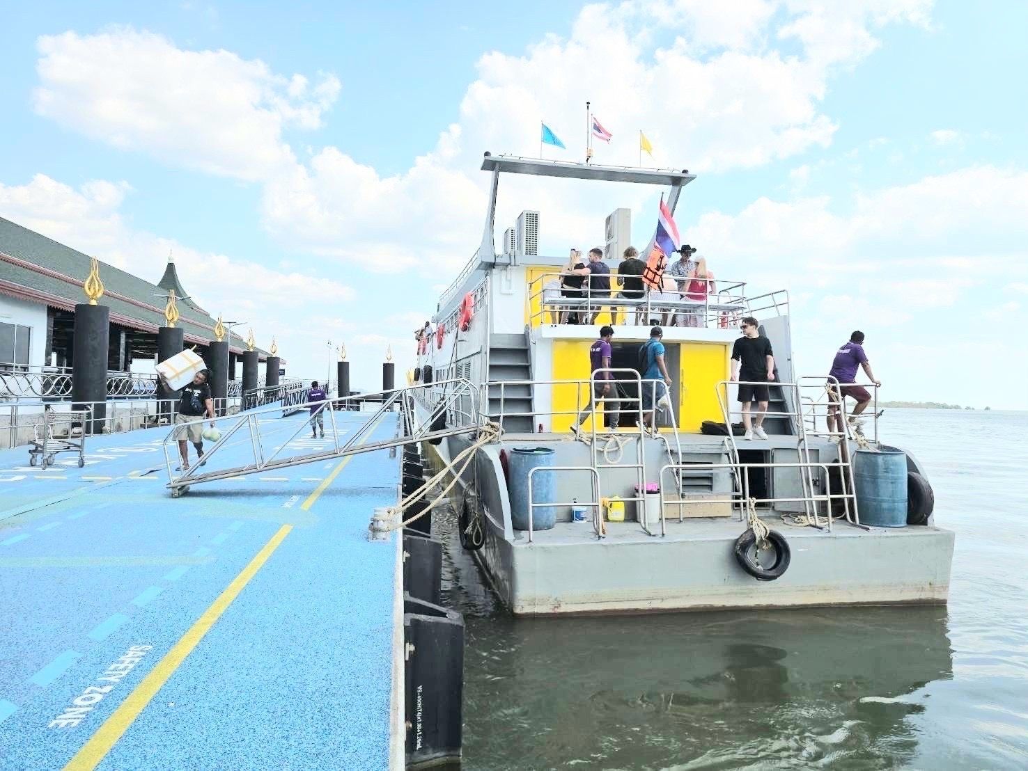 Passageiros a embarcar num ferry branco e amarelo atracado junto a um cais de betão azul sob céu nublado, na Tailândia