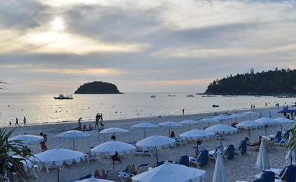 Praia com chapéus de sol brancos, visitantes, barcos e ilhas arborizadas sob céu nublado ao anoitecer