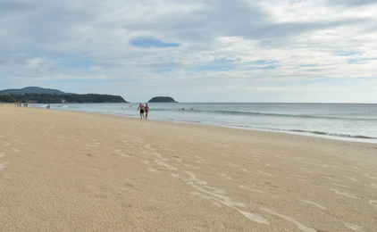 Praia de areia com águas turquesa calmas, ilhas ao longe e algumas pessoas na água junto à margem