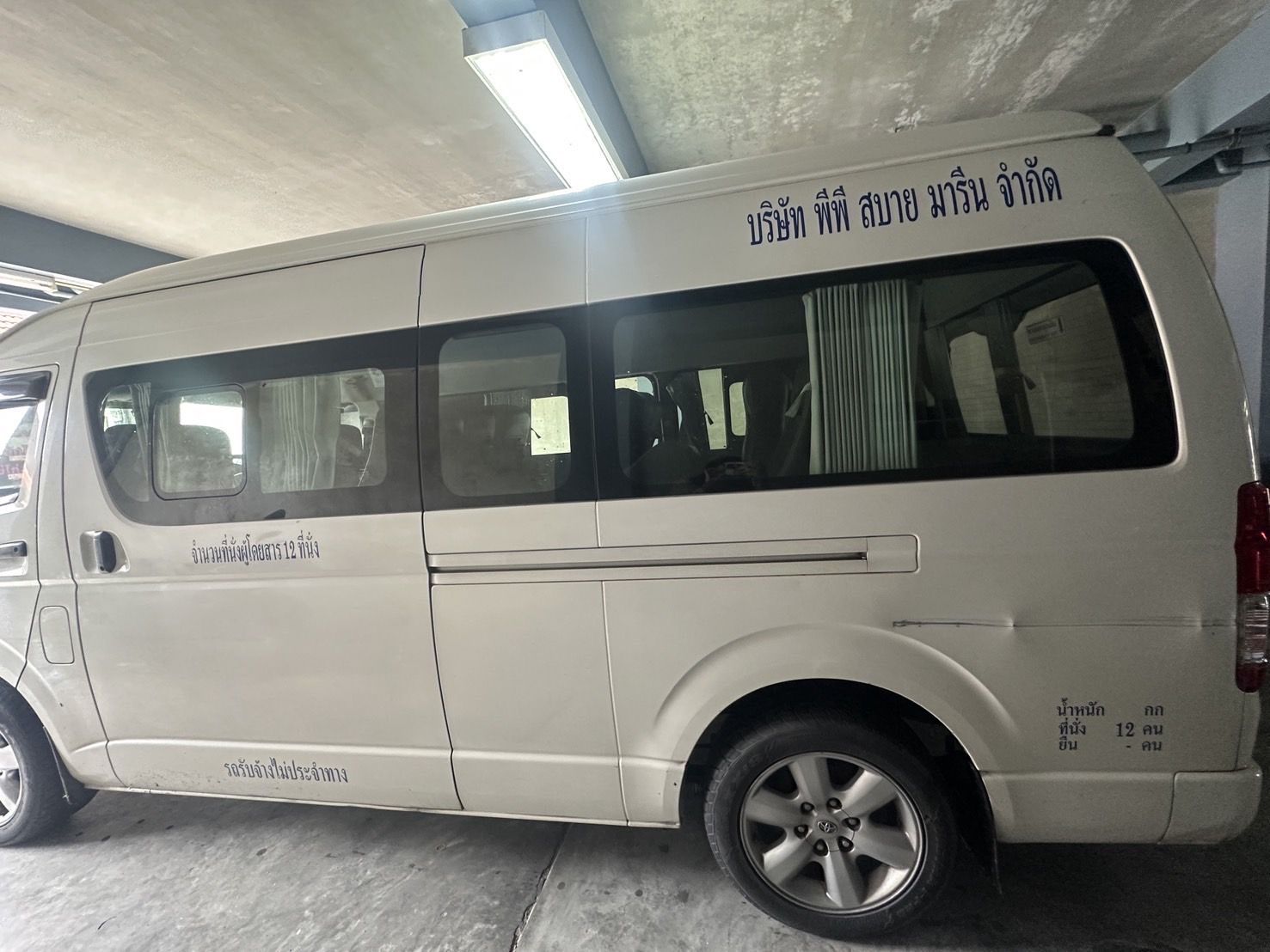 Carrinha Toyota Hiace branca de 12 lugares com letras de empresa tailandesa estacionada sob um abrigo de betão, na Tailândia