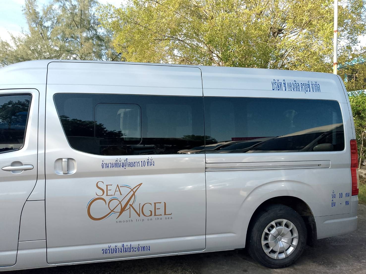 Carrinha prateada de 10 lugares com marca "Sea Angel" e letras tailandesas estacionada sob árvores num ponto de transferência