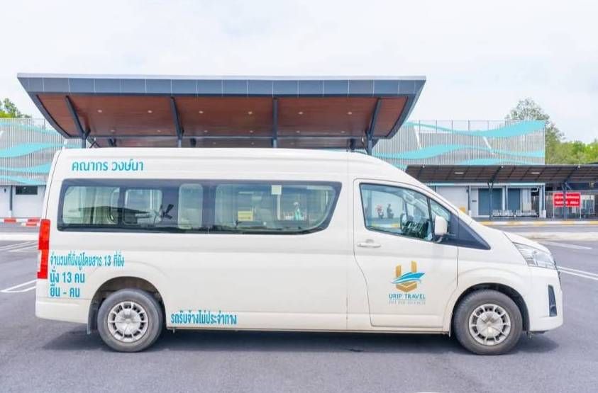 Furgoneta branca de 13 lugares com letras tailandesas estacionada sob o alpendre coberto de um terminal em asfalto cinzento