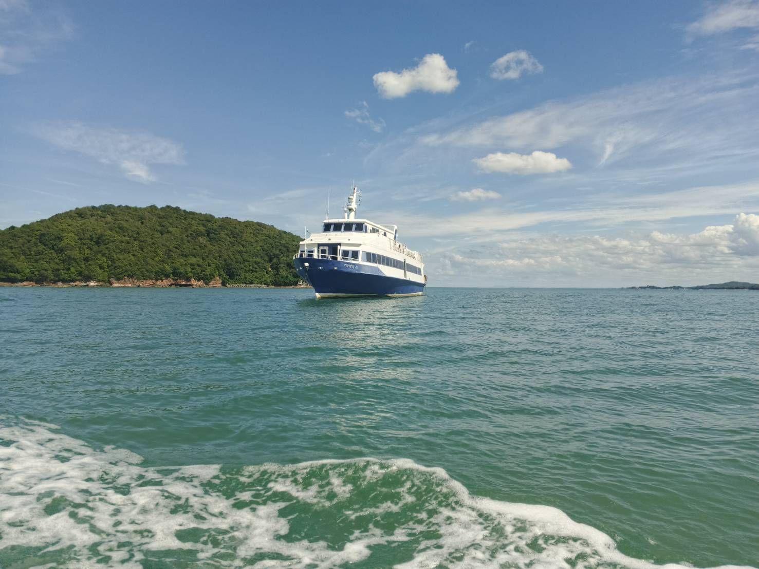 Ferry de passageiros azul e branco fundeado em mar turquesa junto a uma ilha verde arborizada sob céu azul
