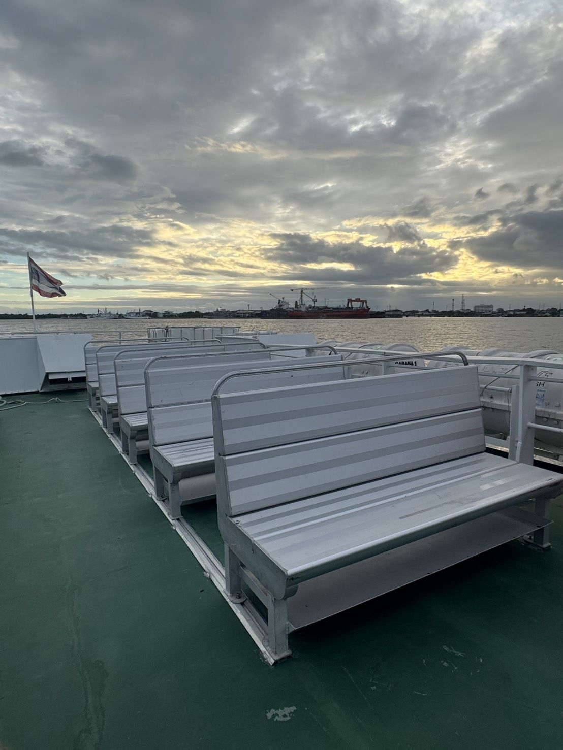 Bancos de alumínio vazios no convés verde ao ar livre de um ferry, bandeira tailandesa, navio de carga no rio ao anoitecer