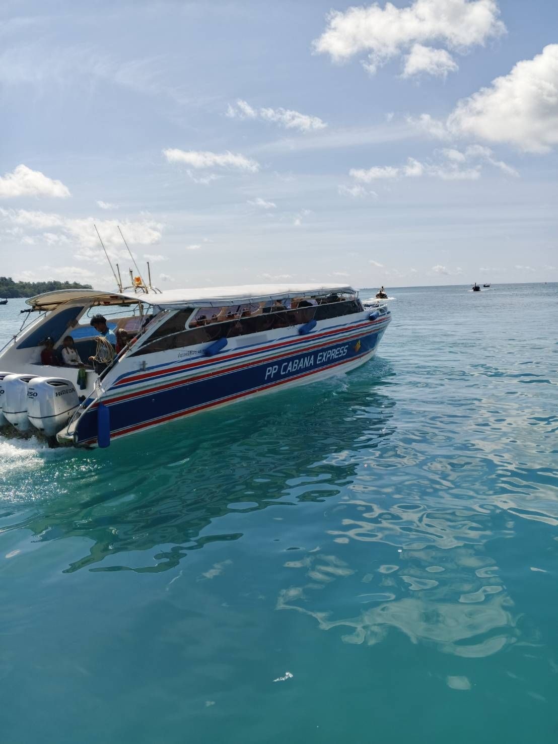 Lancha azul e branca "PP Cabana Express" com passageiros a atravessar água tropical turquesa sob céu parcialmente nublado