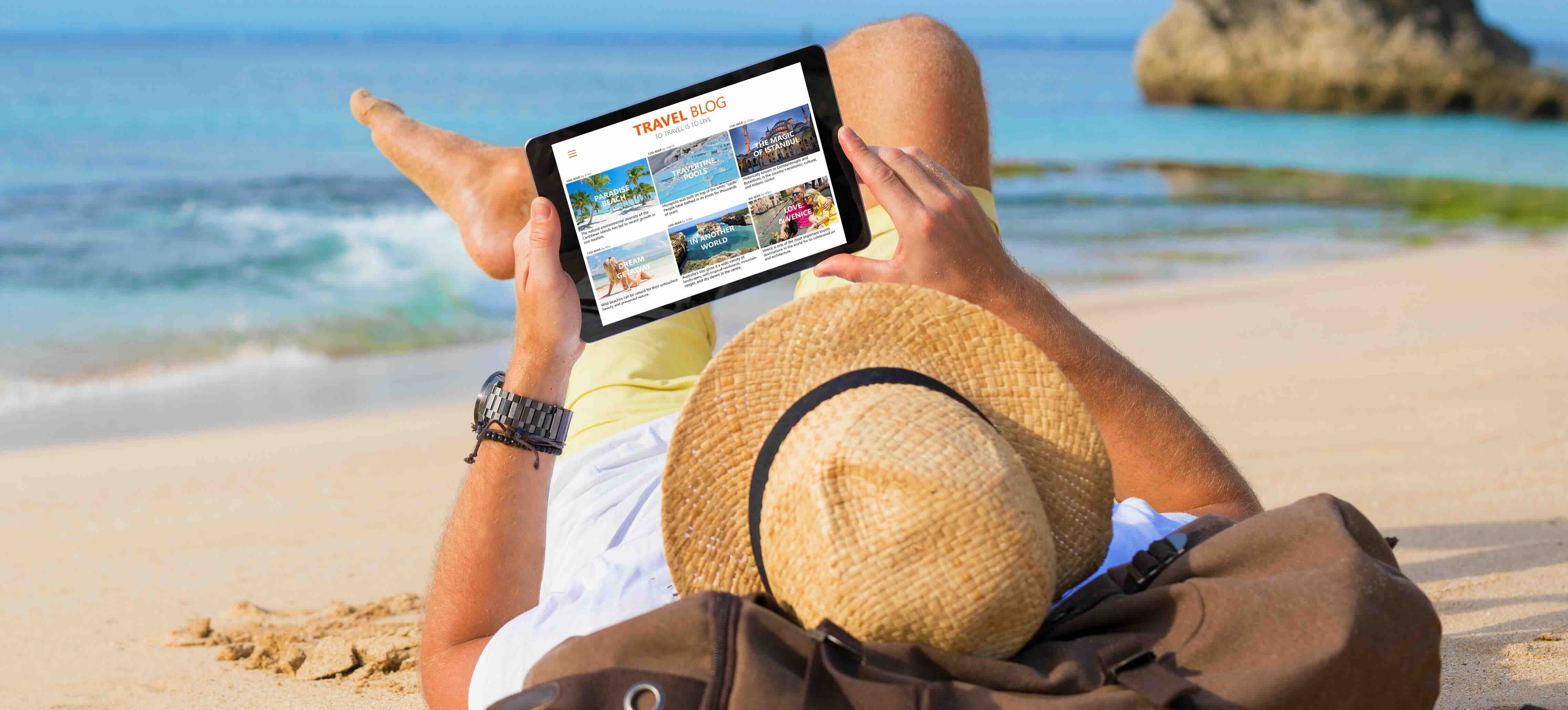 Homem deitado numa praia de areia, com chapéu de palha e camisa amarela, a ler um blogue de viagens num tablet