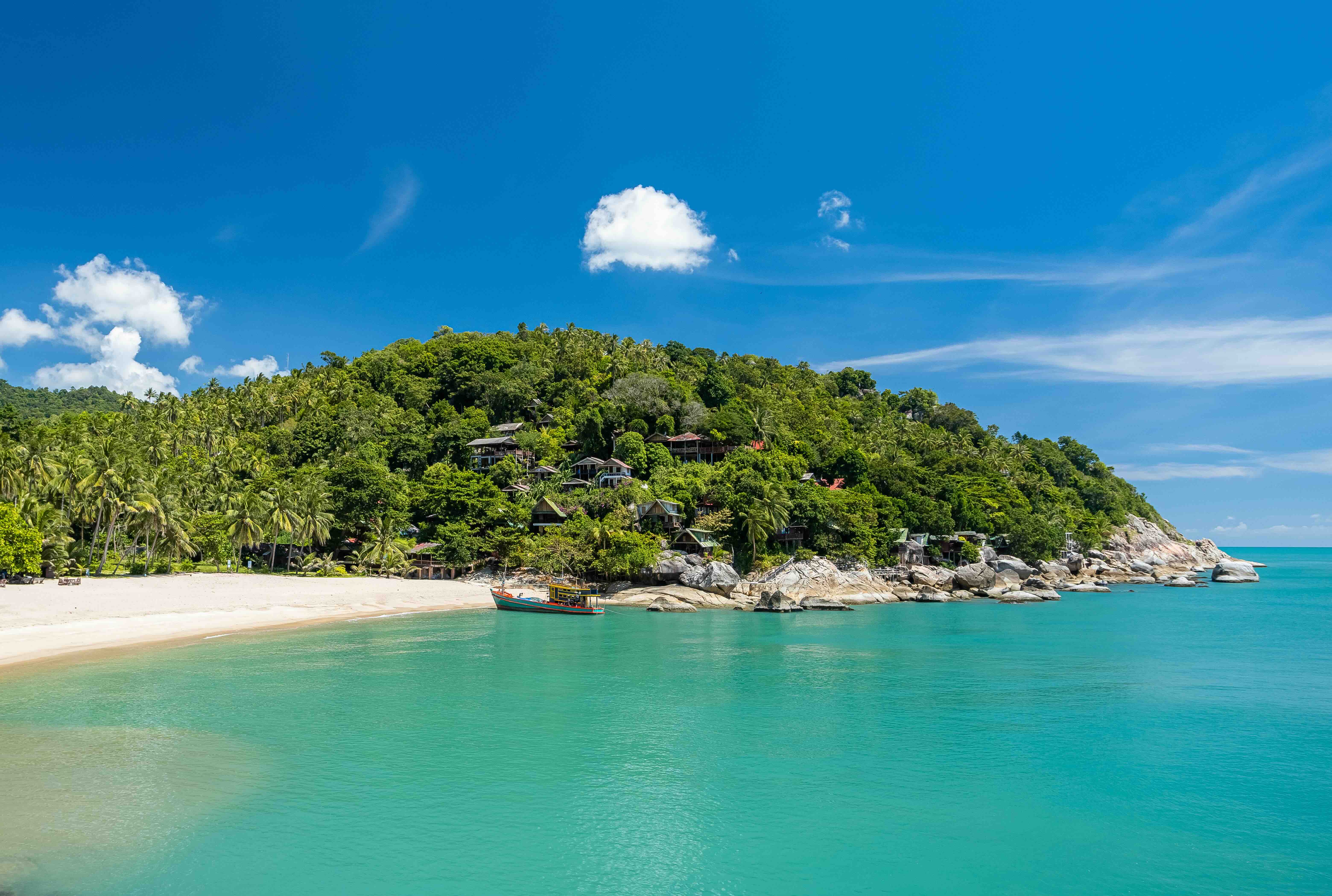 Praia de ilha tropical com água turquesa, areia, vegetação exuberante, falésias rochosas e barco de madeira