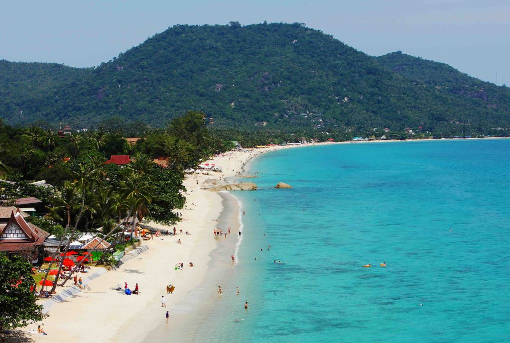 Praia turquesa com areia branca, palmeiras, colinas florestadas e banhistas a nadar em água cristalina