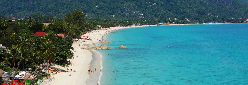 Baía turquesa com praia de areia branca, palmeiras e edifícios residenciais na encosta