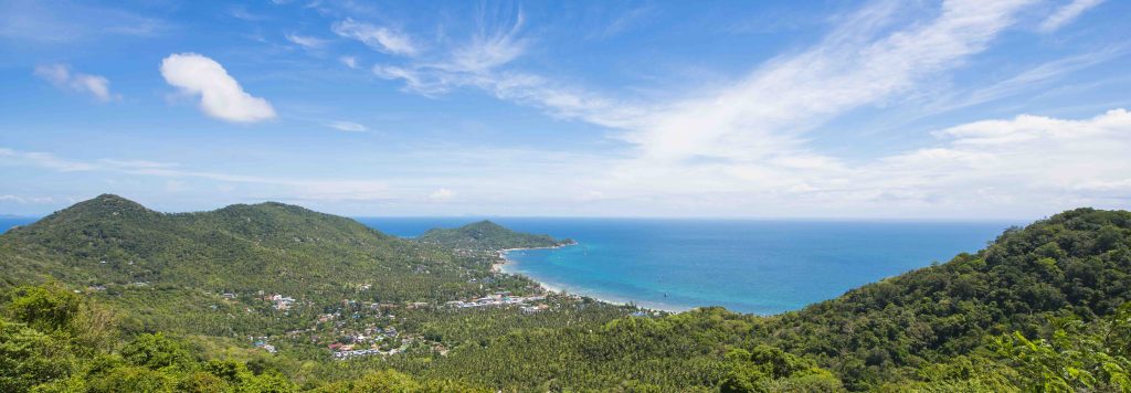 Collines verdoyantes descendant vers une eau turquoise et une plage de sable avec village côtier, ciel bleu