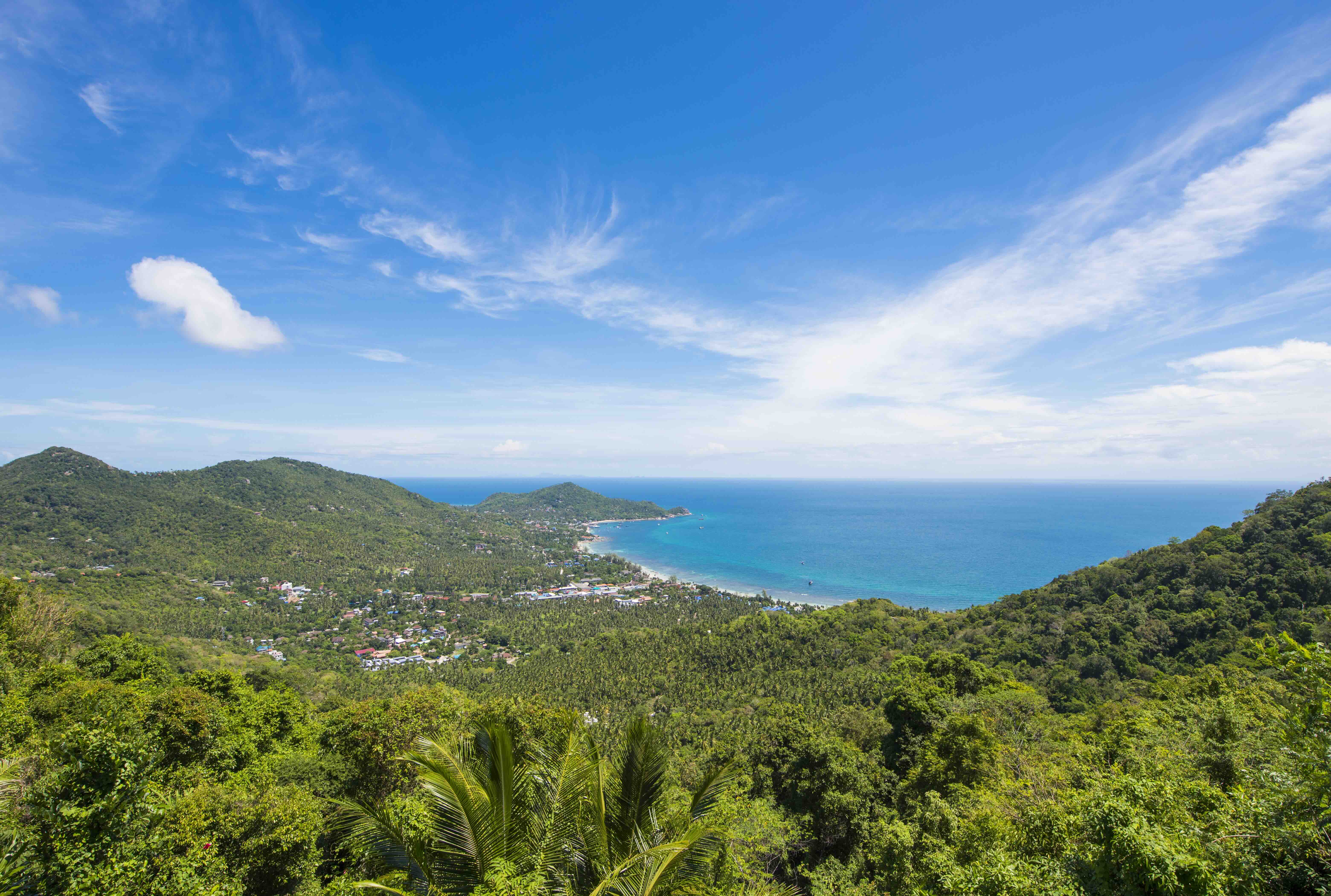 Vista aérea de colinas verdejantes que descem até uma baía turquesa com praia de areia e povoação costeira
