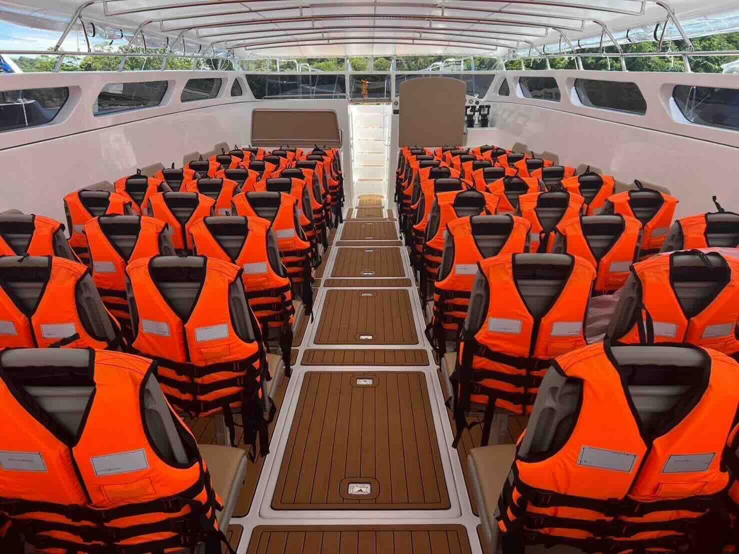Cabina de passageiros vazia de uma lancha com filas de assentos cobertos por coletes salva-vidas laranja e corredor com aspeto de teca