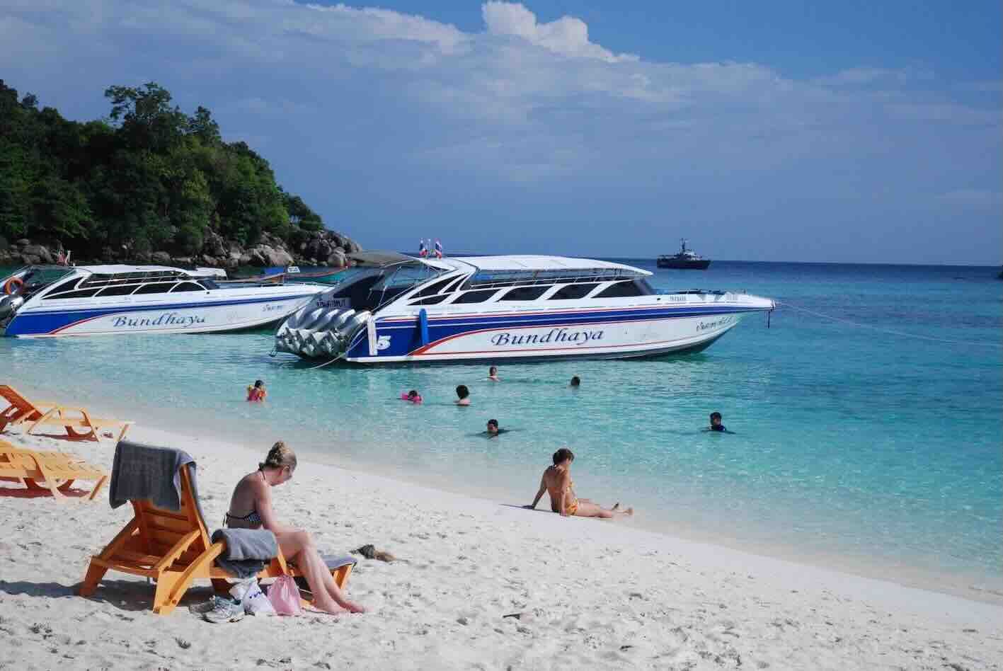 Lanchas com a inscrição "Bundhaya" ancoradas numa praia de areia branca enquanto banhistas caminham em água turquesa cristalina