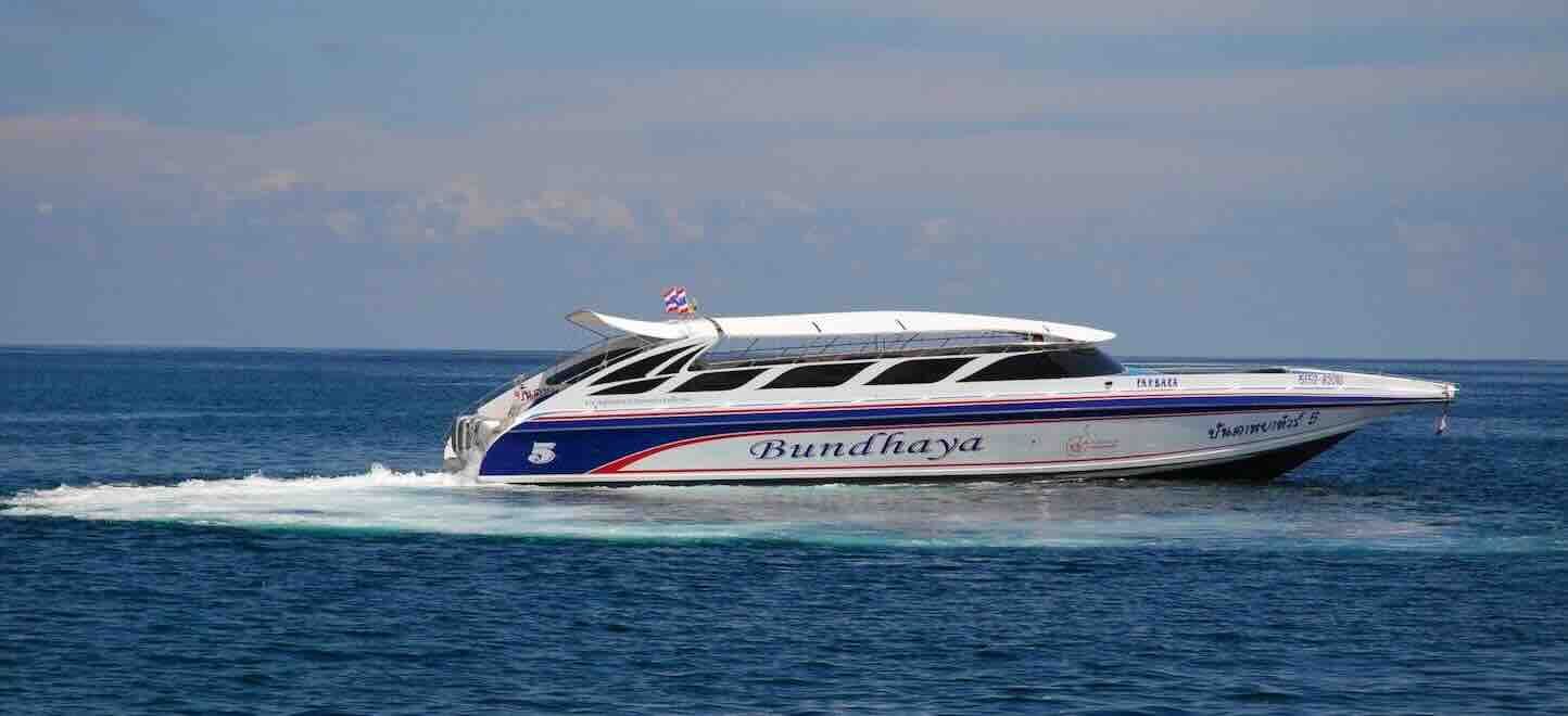 Lancha Bundhaya branca com faixas azuis e vermelhas a navegar em mar azul aberto sob céu enevoado