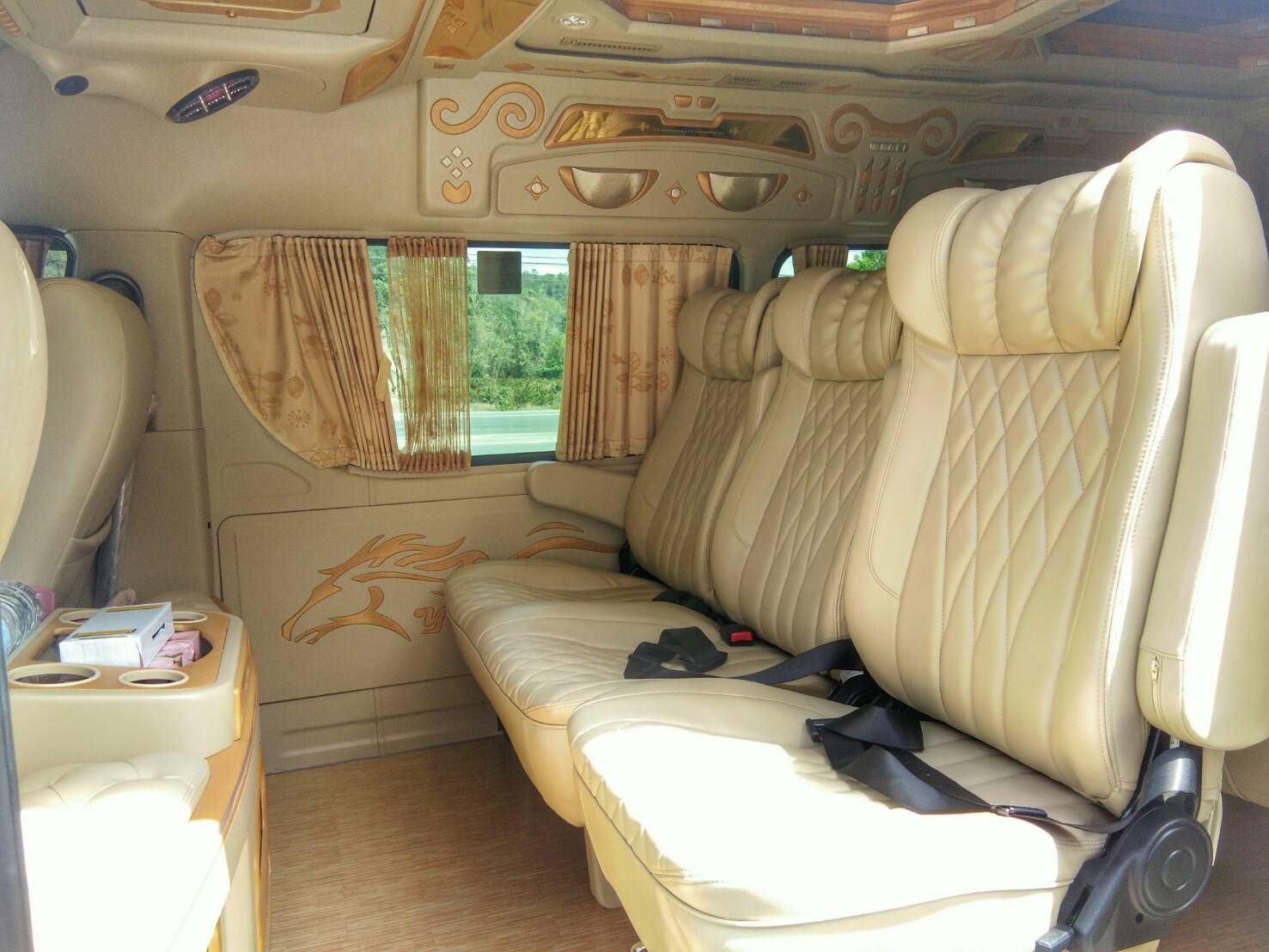 Interior de uma minivan VIP com assentos reclináveis de couro creme, acabamentos dourados, janelas com cortinas e porta-copos