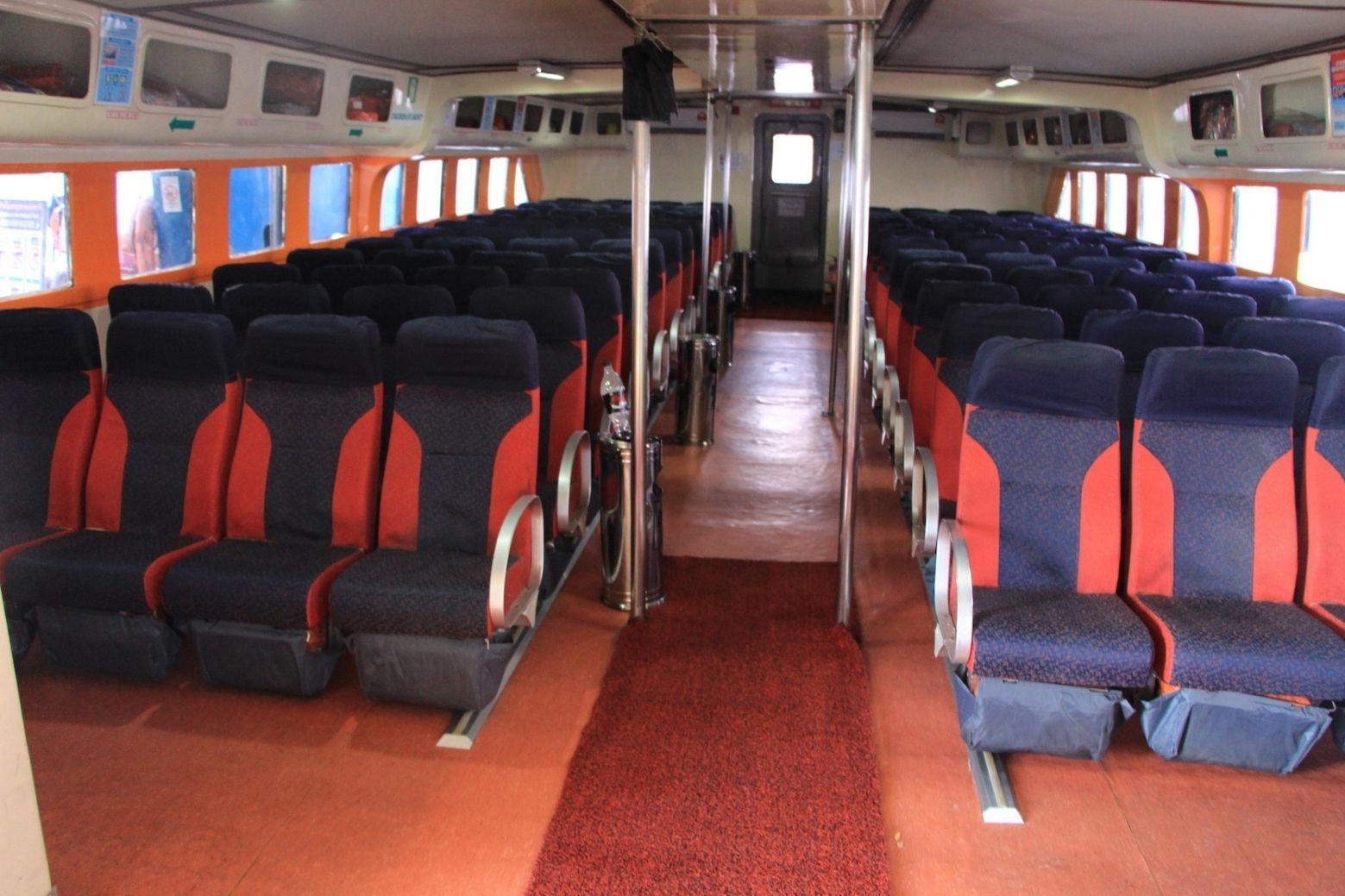 Cabina de passageiros vazia de um ferry rápido com filas de assentos vermelhos e azul-marinho, paredes laranja e corredor central