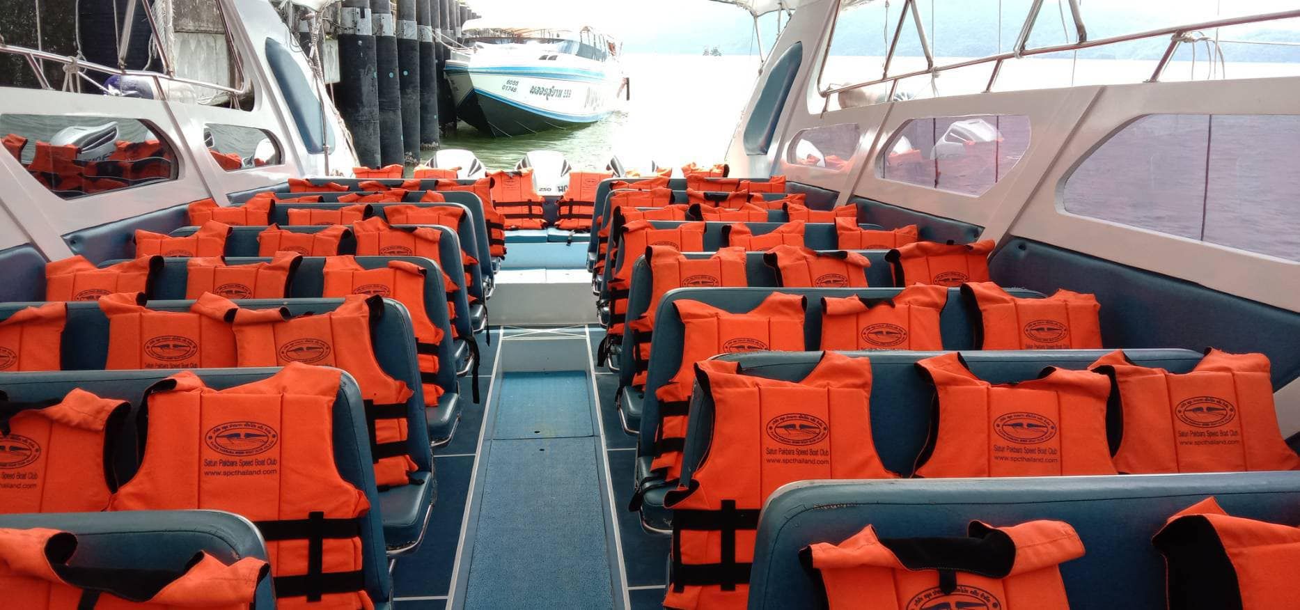 Cabina de passageiros de lancha com filas de assentos azuis cobertos por coletes salva-vidas laranja e um barco atracado ao cais atrás