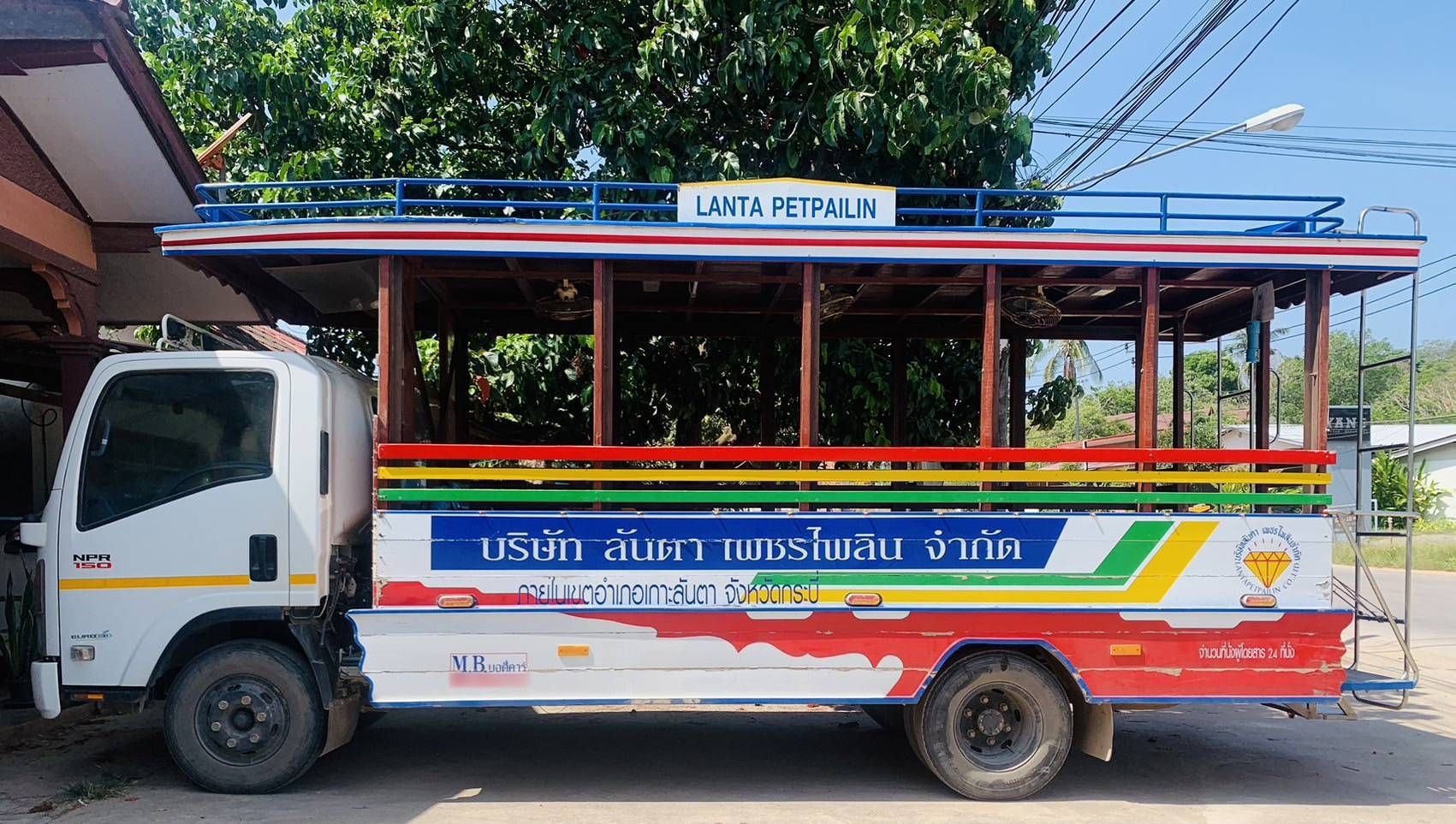 Songthaew de laterais abertas pintado de vermelho, azul e branco com letras tailandesas e placa "Lanta Petpailin" no teto, estacionado na estrada