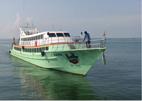 Ferry de passageiros tailandês verde-menta fundeado em mar calmo, com um tripulante à proa junto à bandeira tailandesa