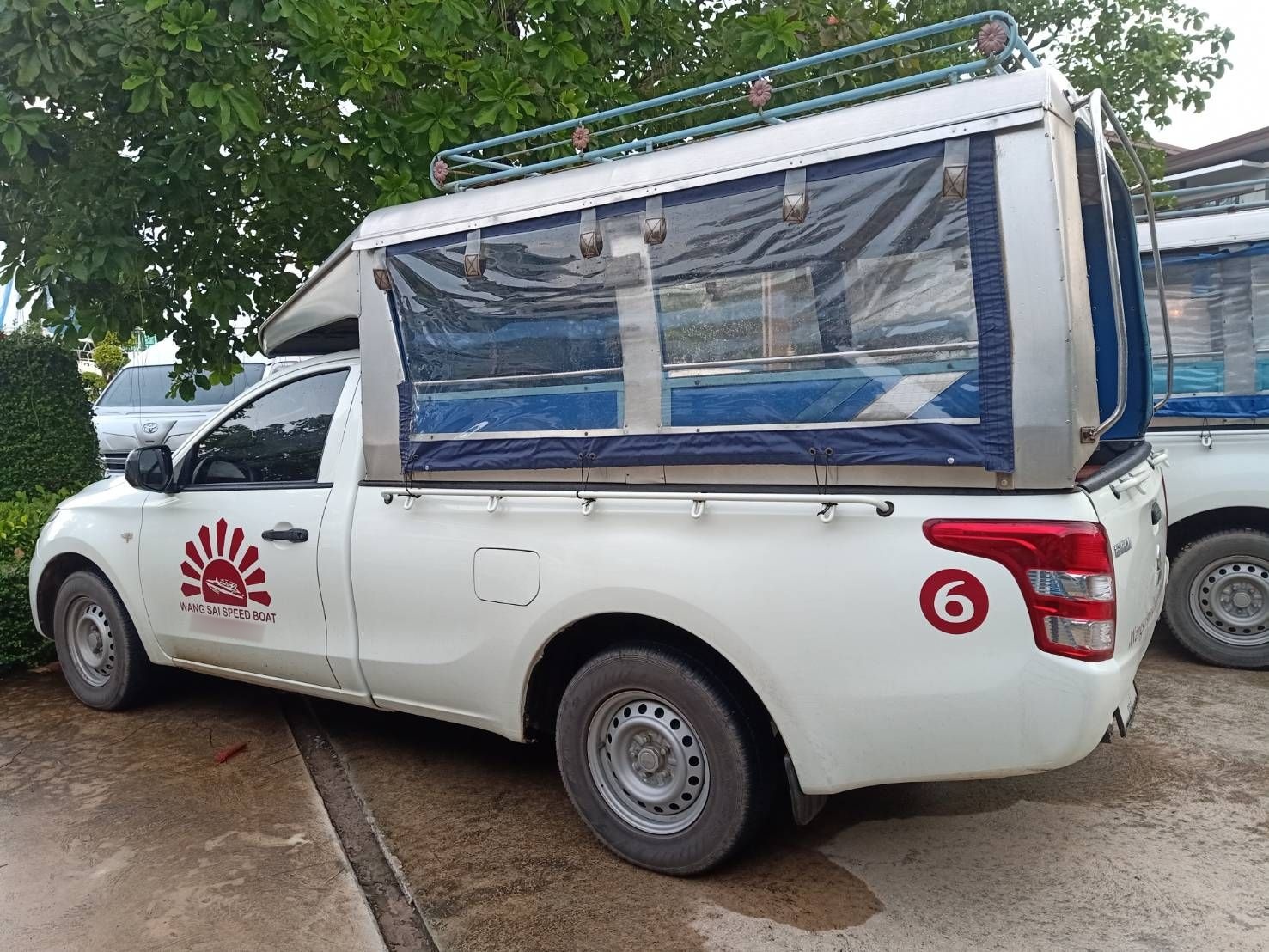 Pick-up songthaew branca com banco de passageiros coberto, logótipo de transferência de barco em sol vermelho e o número 6, estacionada em piso de cimento
