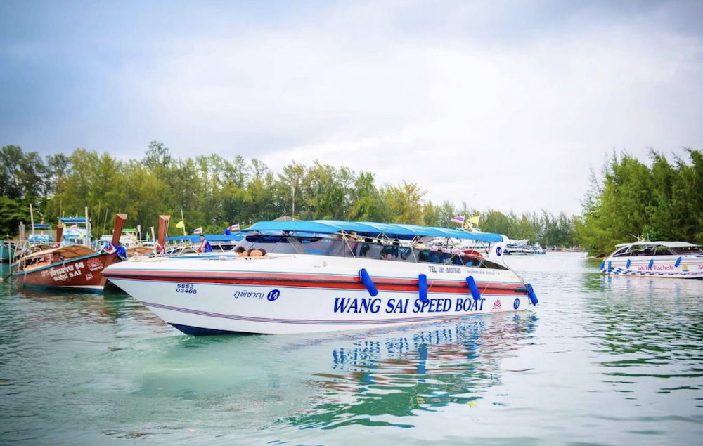 White Wang Sai speed boat with blue canopy moored in calm green water beside wooden longtail boats at a Thai pier