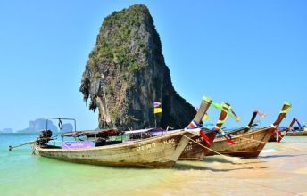Barcos longtail de madeira ancorados numa praia turquesa de água rasa sob uma imponente formação calcária, costa ao estilo de Railay, Tailândia