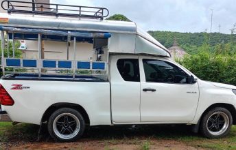 Pick-up branca convertida em songthaew com bancos azuis e bagageira no tejadilho, na relva junto a colinas verdes